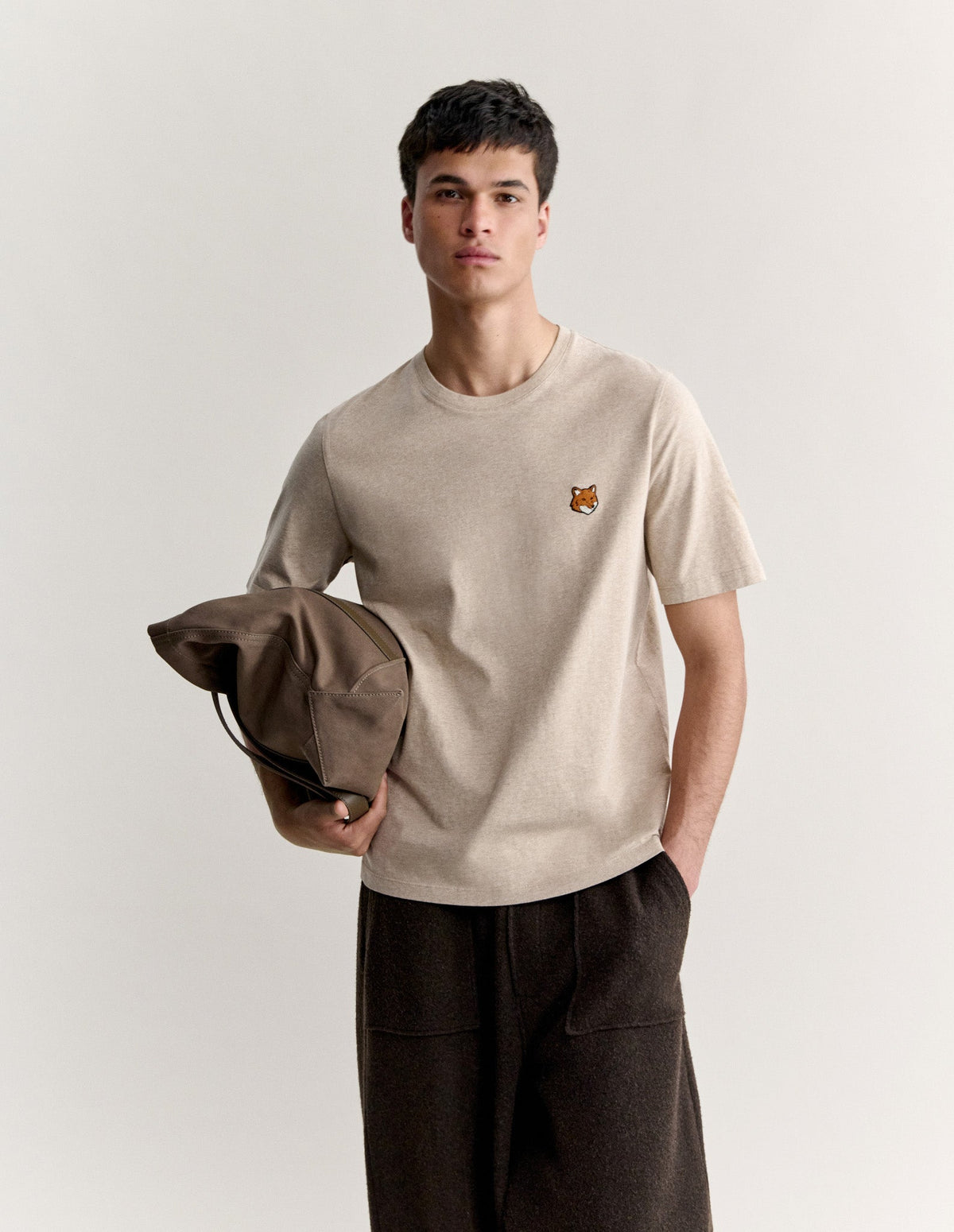 Hombre con la camiseta Maison Kitsuné Fox Head beige y pantalón oscuro, sujetando una prenda, vista de cuerpo entero