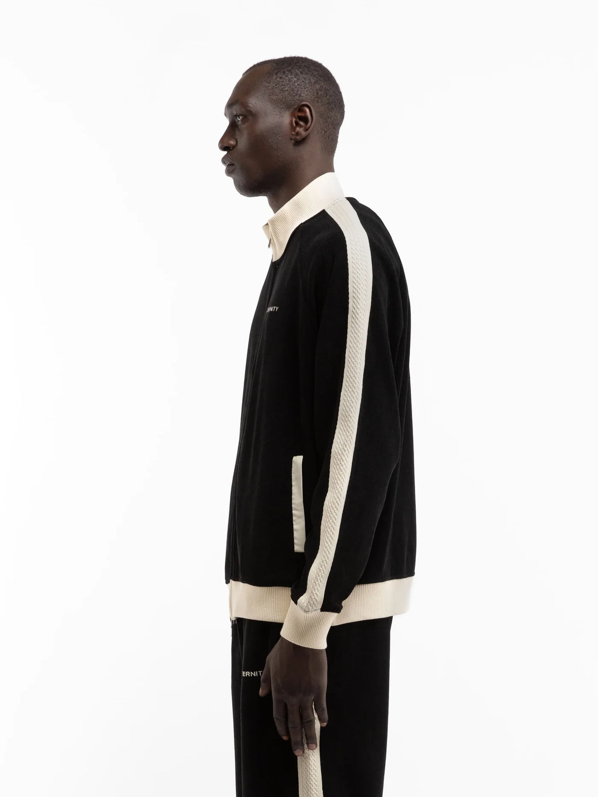 Hombre de perfil con la chaqueta Reternity Studio Knit Track negra con ribetes crema, cuerpo entero