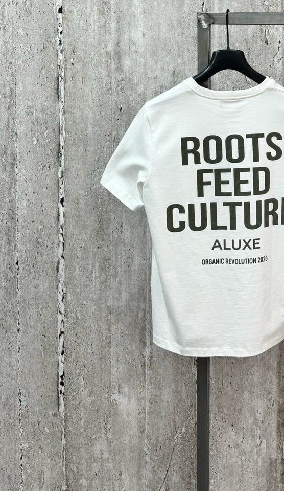 Camiseta blanca colgada con estampado Roots Feed Culture de ALUXE sobre pared de hormigón gris
