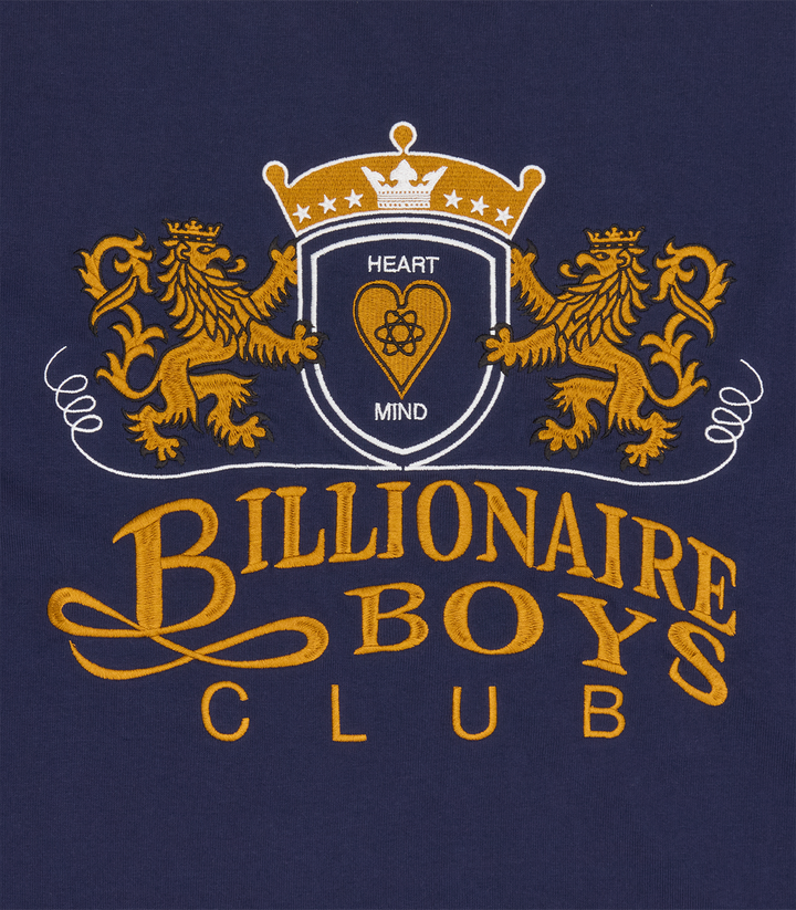 Detalle del escudo con dos leones bordado, corona y BILLIONAIRE BOYS CLUB en dorado sobre la camiseta azul marino
