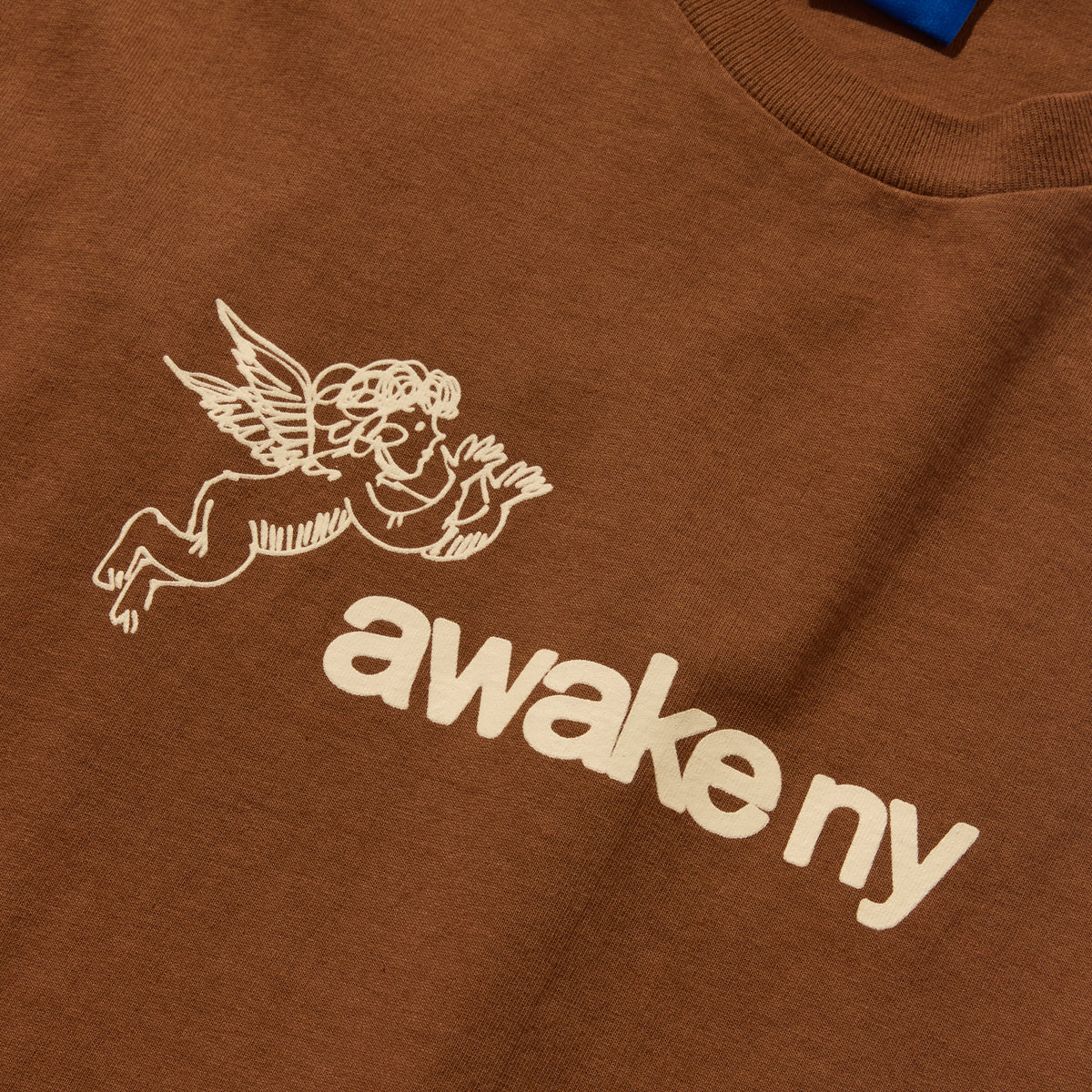 Detalle del querubín alado y el logo awake ny bordado en crema sobre la camiseta marrón