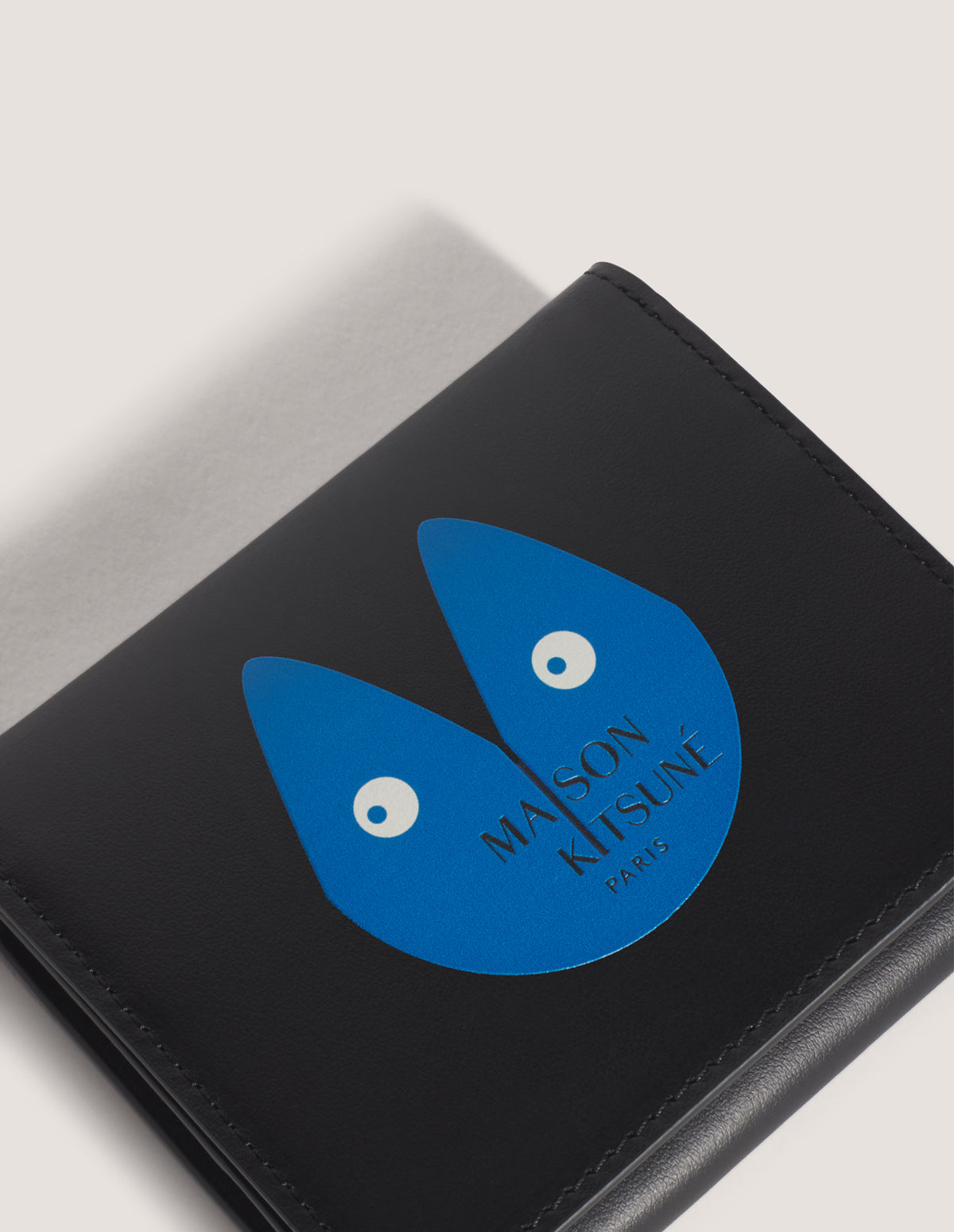 Detalle del zorro en azul sobre la cartera Maison Kitsuné Fortune Fox negra, vista en tres cuartos