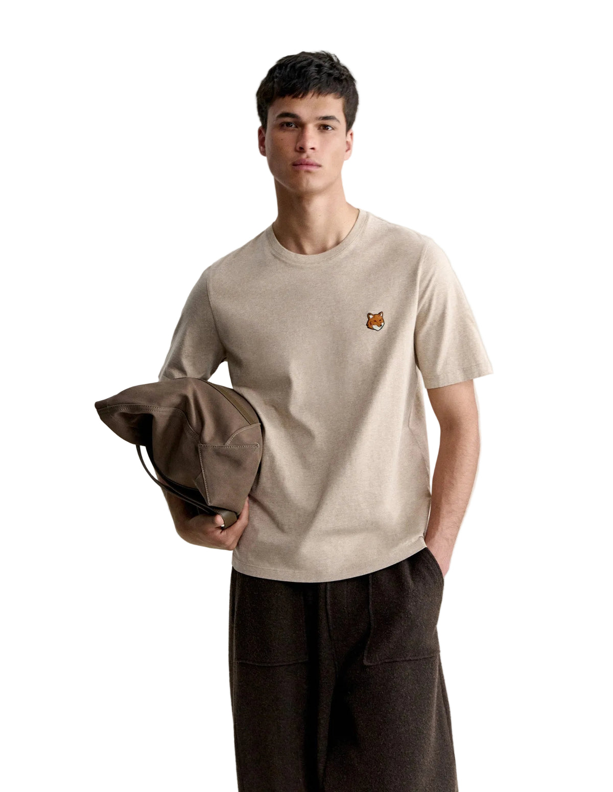 Hombre con la camiseta Maison Kitsuné Fox Head beige y pantalón oscuro, sujetando una prenda, vista de cuerpo entero